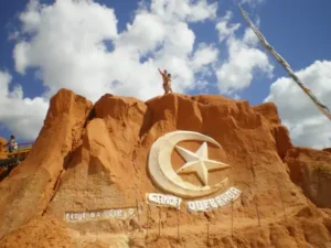 A foto mostra o icônico símbolo de Canoa Quebrada, uma lua crescente com uma estrela em seu interior, esculpido na face de uma falésia avermelhada. A falésia, de textura arenosa e irregular, destaca-se contra o céu azul e parcialmente nublado. Duas pessoas estão no topo da falésia, com os braços abertos, possivelmente celebrando a vista. A inscrição "Canoa Quebrada" aparece logo abaixo do símbolo da lua e estrela, reforçando a identidade do local. A cor vibrante da falésia e a imponência do símbolo criam uma imagem marcante da paisagem de Canoa Quebrada, um dos passeios diários oferecidos pelo Refúgio Hostel Fortaleza & Pousada.